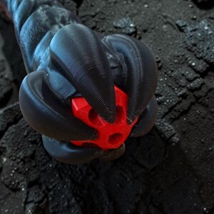 May include: A close-up of a black, textured object with a red, geometric shape at its center. The object appears to be made of a 3D-printed material, with a dark, rough background.