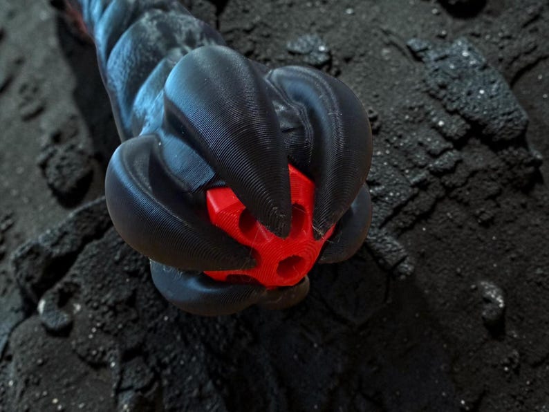 May include: A close-up of a black, textured object with a red, geometric shape at its center. The object appears to be made of a 3D-printed material, with a dark, rough background.