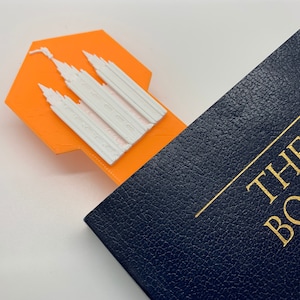 May include: Orange bookmark with a white 3D printed temple design. The bookmark is placed on a blue book with gold lettering that reads "THE BOOK OF"