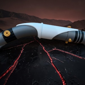 K&ouml;nnte beinhalten: Ein futuristisches, gebogenes Lichtschwert-Requisit mit einem grau-wei&szlig;en K&ouml;rper, akzentuiert mit schwarzen Streifen und gelben kreisf&ouml;rmigen Details. Das Requisit befindet sich auf einer dunklen, strukturierten Oberfl&auml;che mit roten Adern und einem Hintergrund aus Bergen unter einem dunklen Himmel.