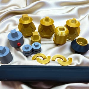 May include: An assortment of 3D-printed objects in various shapes and colors. The collection includes gold-colored hexagonal containers with square lids, gray cylinders, and a long, rectangular dark blue object. Also, there are gold-colored curved shapes.
