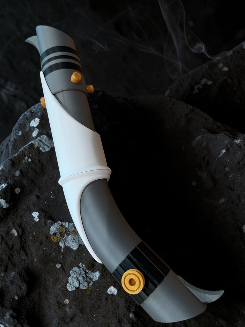 K&ouml;nnte beinhalten: Ein graues und wei&szlig;es Lichtschwert-Requisit mit schwarzen Akzenten und goldfarbenen Details. Die Requisite hat ein gebogenes Design und befindet sich vor einem dunklen, strukturierten Hintergrund. Das Lichtschwert hat eine elegante, futuristische &Auml;sthetik.