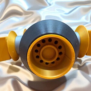 May include: Close-up of a 3D-printed object with a black central component and yellow-orange accents. The central piece has a series of small holes. The object is set against a shiny, silvery background.
