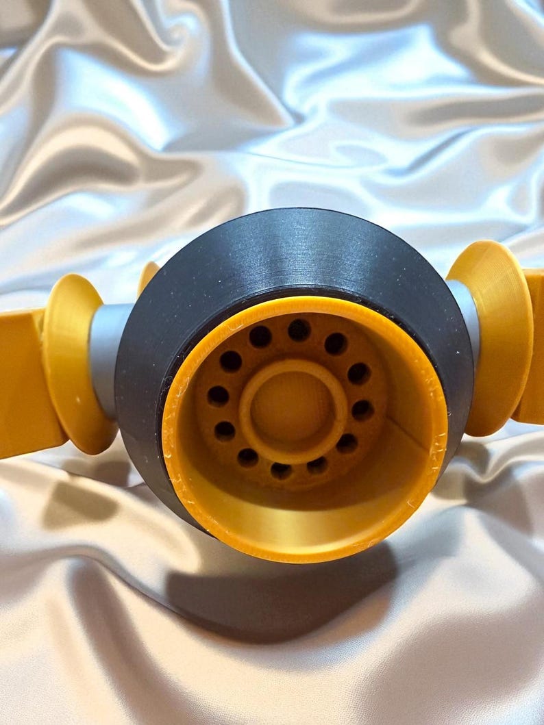 May include: Close-up of a 3D-printed object with a black central component and yellow-orange accents. The central piece has a series of small holes. The object is set against a shiny, silvery background.