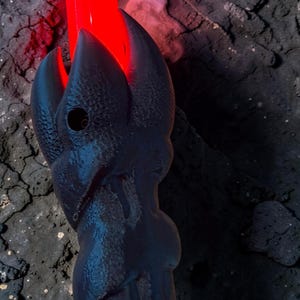 May include: A black, stylized lightsaber hilt with a red blade emitting a bright glow. The hilt has a textured, claw-like design. Smoke-like vapor rises from the blade, creating a dramatic effect. The background is dark and textured.