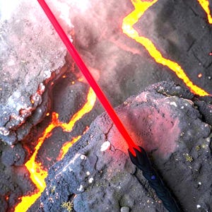 May include: A red lightsaber with a black handle, resting on a dark, textured surface with a lava-like background. The lightsaber's blade glows brightly, contrasting with the dark handle and the fiery backdrop.