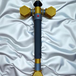 May include: A 3D-printed hammer with a dark blue handle and gold-colored hexagonal heads. The hammer features a red button and a black top. The object is set against a white, satin-like background.