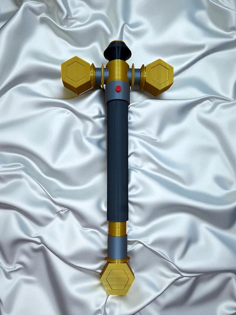 May include: A 3D-printed hammer with a dark blue handle and gold-colored hexagonal heads. The hammer features a red button and a black top. The object is set against a white, satin-like background.