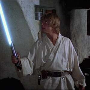 May include: A man in a white robe holds a blue lightsaber in his right hand. He is standing in a dimly lit room with a golden robot standing to his left. A man in a white robe is sitting in the background.