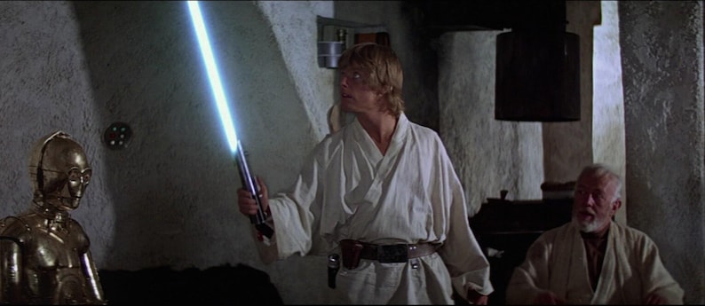 May include: A man in a white robe holds a blue lightsaber in his right hand. He is standing in a dimly lit room with a golden robot standing to his left. A man in a white robe is sitting in the background.