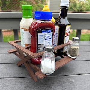 Puede incluir: Un soporte de condimentos de madera marrón con forma de mesa de picnic con cuatro botellas de condimentos y dos saleros y pimenteros. Las botellas incluyen ketchup, aderezo para ensaladas, salsa barbacoa y mostaza.