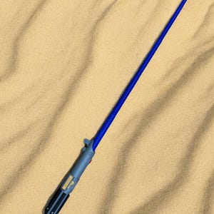 May include: A blue and gray lightsaber toy on a sandy surface.
