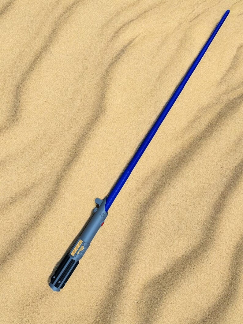 May include: A blue and gray lightsaber toy on a sandy surface.