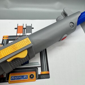 May include: A 3D printed gray lightsaber with a yellow and orange handle. The lightsaber is being printed on a 3D printer with the text "PRINTING...89" displayed on the screen.