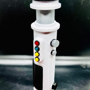 Puede incluir: Un accesorio de sable l&aacute;ser blanco con una franja negra y una serie de botones de colores. Los botones son rojo, verde, azul y amarillo.