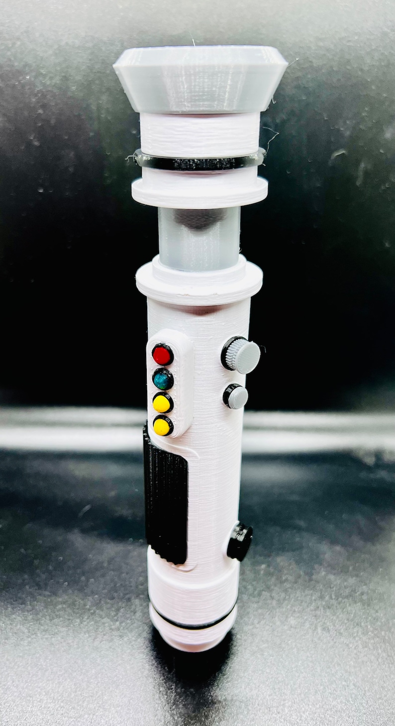 Puede incluir: Un accesorio de sable l&aacute;ser blanco con una franja negra y una serie de botones de colores. Los botones son rojo, verde, azul y amarillo.