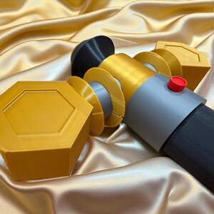 May include: A multi-colored, geometric-shaped object with a black, gray, gold, and red color scheme. The object has a cylindrical shape with a red button on the side. The background is a gold satin fabric.