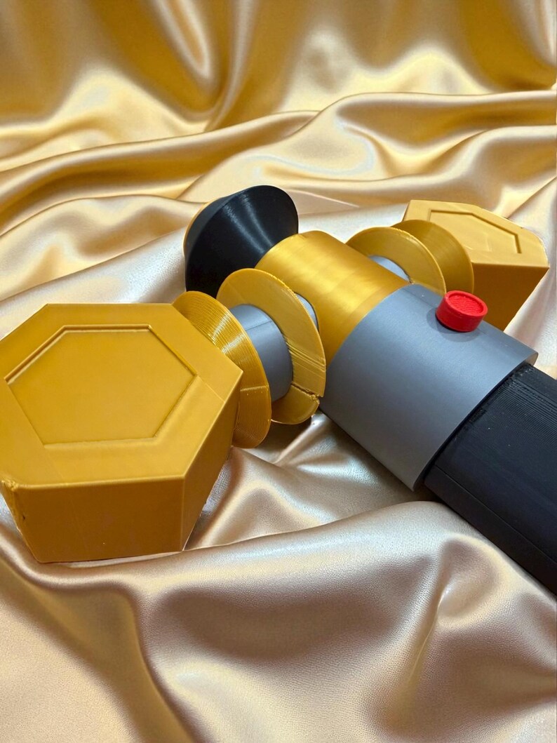 May include: A multi-colored, geometric-shaped object with a black, gray, gold, and red color scheme. The object has a cylindrical shape with a red button on the side. The background is a gold satin fabric.