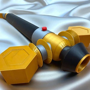 May include: A 3D-printed prop weapon with a black, gold, and gray color scheme. The prop features a cylindrical body with geometric shapes and a red button. The design includes a black handle and gold accents.