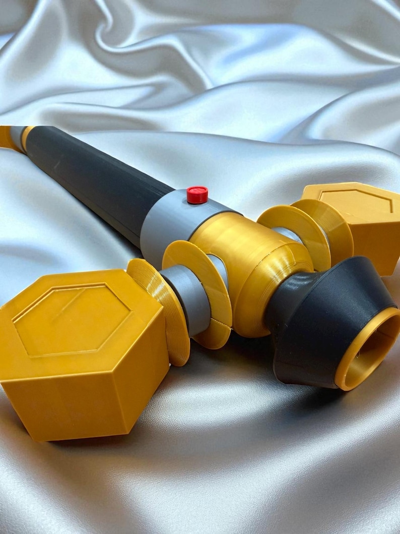 May include: A 3D-printed prop weapon with a black, gold, and gray color scheme. The prop features a cylindrical body with geometric shapes and a red button. The design includes a black handle and gold accents.