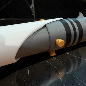 K&ouml;nnte beinhalten: Ein grauer und wei&szlig;er Lichtschwertgriff mit schwarzen Streifen und orangefarbenen Akzenten. Der Griff hat ein zylindrisches Design mit einem gebogenen wei&szlig;en Abschnitt. Zwei orangefarbene Kn&ouml;pfe sind sichtbar.