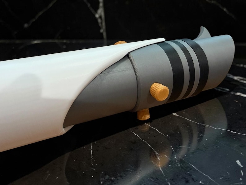 K&ouml;nnte beinhalten: Ein grauer und wei&szlig;er Lichtschwertgriff mit schwarzen Streifen und orangefarbenen Akzenten. Der Griff hat ein zylindrisches Design mit einem gebogenen wei&szlig;en Abschnitt. Zwei orangefarbene Kn&ouml;pfe sind sichtbar.