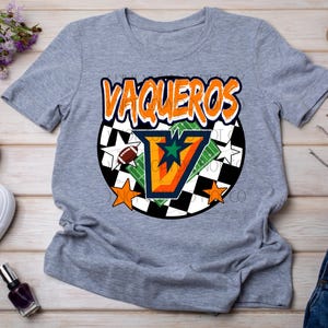 May include: A gray t-shirt with the word "VAQUEROS" in orange and white lettering. The shirt features a football, stars, and a "V" design. The shirt is paired with white sneakers, sunglasses, and denim shorts.