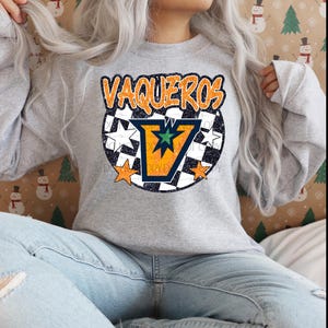 May include: A grey sweatshirt featuring the word "VAQUEROS" in orange. The design includes an orange and blue "V" with a green star, surrounded by white stars and a black and white chequered pattern. A casual top.