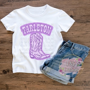 Könnte beinhalten: Ein weißes T-Shirt mit einem lila Aufdruck, der "TARLETON" und einen lila Cowboy-Stiefel zeigt. Dazu eine Jeans-Shorts. Das Bild ist auf einer Holzoberfläche platziert.