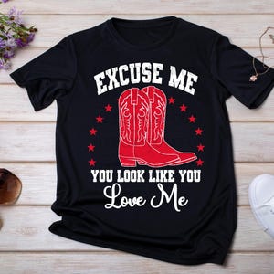 May include: Black t-shirt with white text that reads "Excuse me you look like you love me". The text is surrounded by red stars. There are two red cowboy boots with white stitching on the shirt.