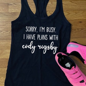 May include: A navy blue racerback tank top with white text that reads "SORRY, I'M BUSY. I HAVE PLANS WITH cody rigsby." Bright pink cycling shoes are next to the tank top.