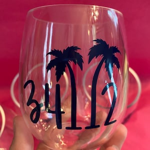 May include: Clear wine glass with a black palm tree design and the numbers "3412" in a script font. The glass is held against a bright pink background, with other glasses visible in the background.