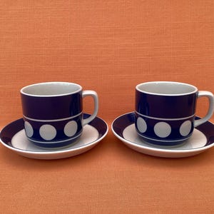 May include: Two matching coffee cups and saucers. The cups and saucers are white with a navy blue border. The cups have a navy blue body with three white polka dots. The background is a solid orange color.