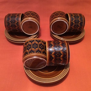 Op de afbeelding: Zes bruine keramische koffiekopjes en schotels. De kopjes hebben een herhalend zwart en bruin geometrisch patroon. De schotels zijn rond met concentrische ringen. De set staat op een oranje oppervlak.