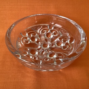 Peut inclure: Un bol en verre transparent avec un motif floral décoratif gravé au fond. Le bol a une forme arrondie et repose sur une surface orange. Le verre est transparent, laissant passer la lumière.