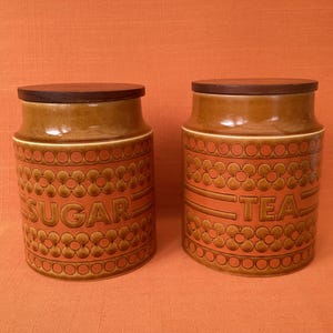 May include: Two cylindrical ceramic canisters with dark brown wooden lids. The canisters feature a glazed orange and brown finish with a repeating embossed floral pattern. One canister is labeled SUGAR and the other is labeled TEA in relief.