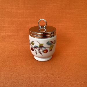 May include: A white ceramic egg coddler with a polished silver metal lid featuring a circular handle. The body of the container is decorated with a botanical pattern of red strawberries, green leaves, and brown stems painted around the exterior.