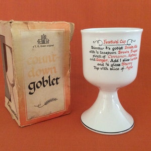 May include: A white ceramic goblet with a black rim and a recipe for a "Festival Cup" printed on the side. The goblet is sitting on a table next to a box that says "Goblet" on it.