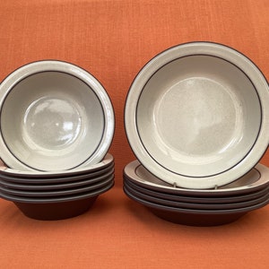 Op de afbeelding: Een set van tien keramische kommen, met een stapel van acht kleinere kommen en twee grotere kommen. De kommen zijn beige met een donkerbruine rand en een donkerbruine binnenkant.