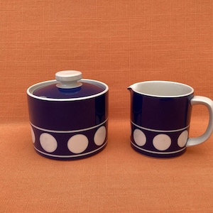 Può includere: Una zuccheriera in ceramica blu navy con coperchio e una lattiera. Entrambi i pezzi hanno un bordo bianco e una fascia bianca con grandi pois bianchi. Il set è su una superficie arancione.