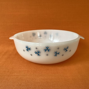 May include: White, round bowl with two handles, decorated with blue starburst patterns. The bowl is made of opaque glass and sits on an orange surface. A vintage kitchenware item.
