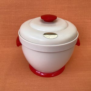 May include: A white and red vintage rice cooker with a rounded shape. The lid has a red knob and the base is also red. The cooker has red handles on the sides and a small metallic label on the front.