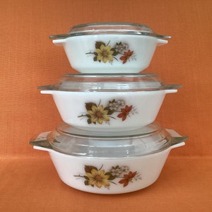 Op de afbeelding: Een stapel van drie witte ovenschaaltjes van melkglas met doorzichtige glazen deksels. Elke schaal heeft een bloemmotief met gele, oranje en bruine bloemen en groene bladeren. De schalen zijn voorzien van geïntegreerde handgrepen aan de zijkant.
