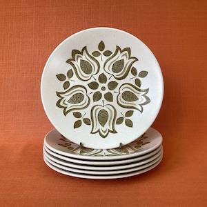 May include: A stack of six white ceramic dinner plates with a green floral design. The plates have a white background with a green floral pattern that features stylized tulips and leaves.