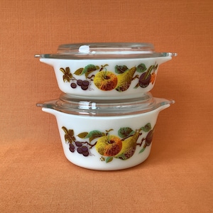 Peut inclure: Deux plats à casserole Pyrex blancs avec couvercles en verre. Chaque plat présente un motif de fruits colorés avec des pommes, des poires et des raisins. Les plats sont empilés les uns sur les autres sur un fond orange.