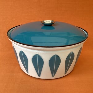 Peut inclure: Une cocotte ronde en émail blanc avec un couvercle assorti bleu sarcelle doté d'un petit bouton métallique. L'extérieur du bol est orné d'un motif répétitif de feuilles verticales bleu sarcelle disposées sur toute la circonférence.