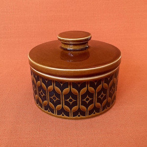 May include: A brown ceramic jar with a lid. The jar has a geometric pattern in black and brown. The lid has a small handle. The jar is sitting on an orange surface.