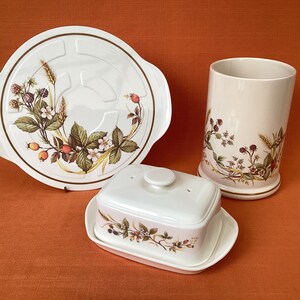 May include: A set of three white ceramic kitchenware pieces with a floral design featuring berries and leaves. The set includes a round serving tray, a butter dish with a lid, and a tall cylindrical jar.