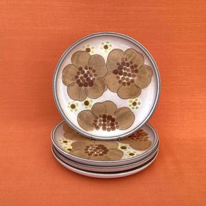 Op de afbeelding: Een stapel van vier keramische borden met een bloemenontwerp. Elk bord heeft een witte basis met bruine en bruine bloemen en een grijze rand. De borden zijn gestapeld op een oranje achtergrond.