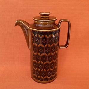 Può includere: Una caffettiera alta in ceramica marrone con coperchio e manico. La caffettiera presenta un motivo ripetuto di forme di foglie stilizzate in un marrone più scuro su uno sfondo marrone più chiaro. Il beccuccio è curvo e il manico è rettangolare.